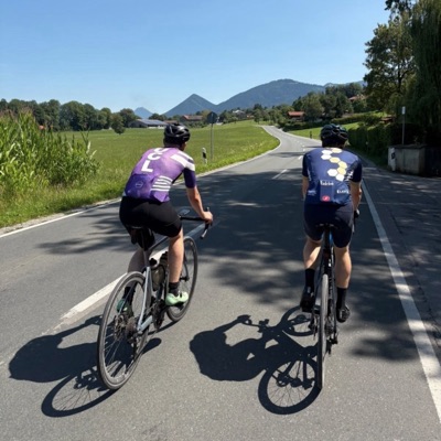 A UCL rider in club kit beside another rider on a valley road