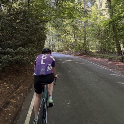 A UCL rider in club kit on a road through woodland, from behind